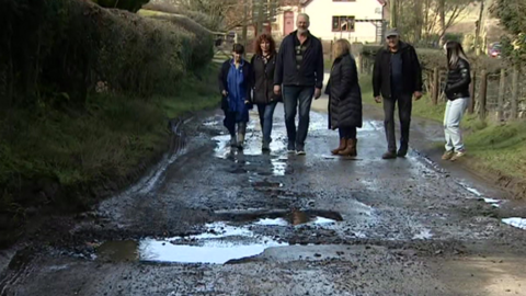 Six Cleeton St Mary residents walk down a potholed-road. A hedge runs along one side and a fence on the other. The residents are in the distance with the potholes in the foreground.