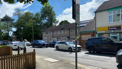 Children queue to cross the road at Middleton Lane as cars go past.