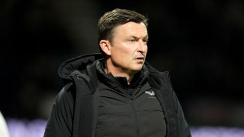 Paul Heckingbottom looks ahead with a focused and thoughtful look on his face. He wears a black zip jacket with a black coat over the top