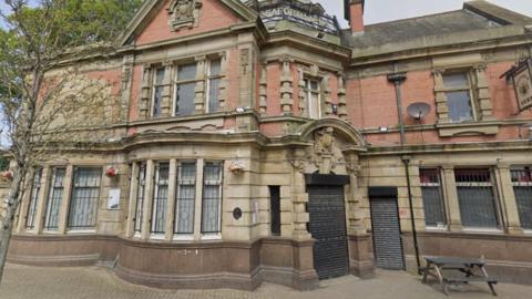 The outside of a red brick and sandstone pub building. A tree grows through a pavement in front of the two-storey building, which can be seen on an overcast today.