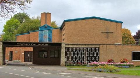 The exterior of Worcester crematorium, a brick building by the side of a road, next to a grassy patch with flowerbeds. The building has a chimney and a stained glass window is visible. A sign above the entrance reads "Worcester Crematorium".