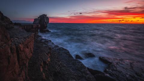 A bright red sunset overlooks a choppy looking sea in front of a rocky coastline