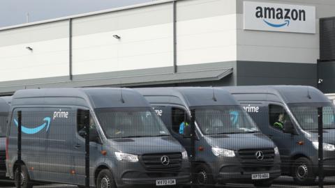 Amazon depot with three vans parked in the foreground with their Amazon livery on a grey background.