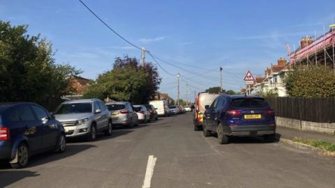 image of Oakfield Road in Bridgwater, Somerset. Cars are parked on either side of the road, and a house in scaffolding can be seen on the right. There is a triangle shaped school sign in the distance.