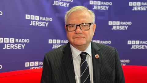 Minister for Children and Families Richard Vilbert is sat in the BBC Radio Jersey studio. He is wearing a suit and tie and is wearing glasses looking straight at the camera. He is sat on a bright red sofa with a purple BBC Radio Jersey branding behind him on the wall.