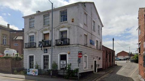 Street view image of a white three-storey building with iron balconies and small trees at the front. The road leads to a car park behind. 