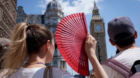 A woman on a crowded pavement near Big Ben cools herself with a bright red handheld fan