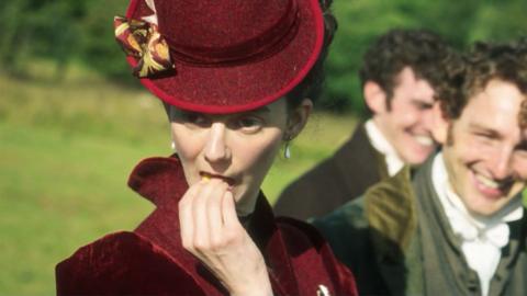 An actress dressed in Georgian clothes eating a primrose covertly, facing away from two men who are laughing behind her