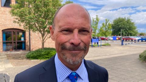 Councillor Adam Kent. He has a partially bald head with a dark grey goatee beard. He is wearing a blue suit and a light blue shirt with a blue star patterned tie. He is stood in courtyard setting with a tree behind him and parked cars in the distance.