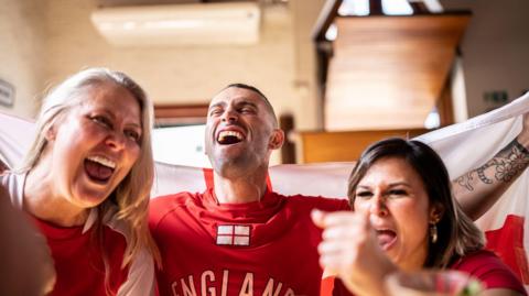 Two women and a man celebrate. They are in a pub wearing England football shirts and holding a flag