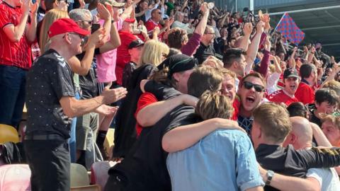 Crowd of people at a football match smiling, shouting and hugging in celebration.