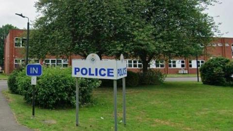 A picture of a police station. There is a white sign in the grounds of the station, that has blue letters spelling police. Behind is a large green tree and behind that is the station.
