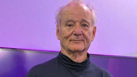 Bill Murray pictured in the BBC Radio WM studio wearing a navy blue turtleneck jumper. He has thin, white hair. He is standing in front of a TV screen which has purple BBC Radio WM branding on it. 