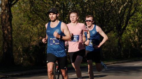 Three men running the Hospice Half-Marathon. Two are wearing blue vests and black shorts, and the third is wearing a pink top and black shorts.