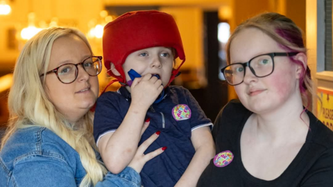 A blonde woman in a denim jacket and a teenage girl with glasses stand on either side of a young boy, both holding him up between them. The boy is wearing a red protective helmet and a navy top, and is holding something to his mouth. They are inside what appears to be a restaurant or function room. 