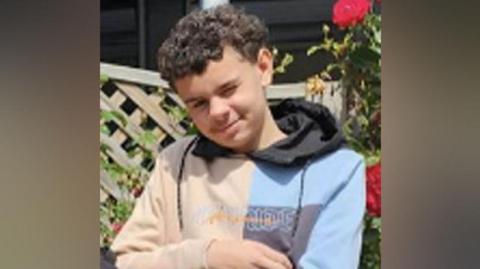 Darrian Williams. He had short curly dark hair and is wearing a blue and beige hoodie. Behind him is a garden trellis and pink roses. He is smiling at the camera.