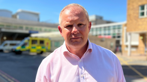 Steve McManus is wearing a pink shirt on a clear day, with the buildings of the hospital trust blurred in the background.