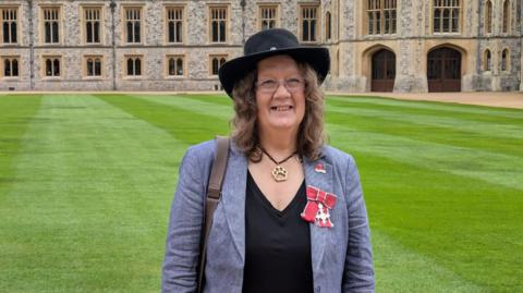 Caroline Gould stands in front of Windsor Castle after being awarded an MBE. She is wearing a grey/blue blazer and a v-neck black t-shirt with a black hat. Her red bow and silver cross MBE is attached to the lapel of her blazer, and she is wearing a gold paw print pendant on a black chain. She is a middle-aged woman with light brown hair and clear-framed glasses, and is smiling.