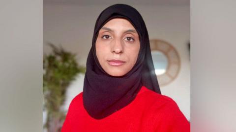 A woman in a red jumper. She has brown eyes and is wearing a black hijab covering her hair.