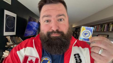 An image of Nikola wearing a Team Canada jacket with a Bosnia jersey underneath it. He has a long dark beard and a nose ring. He is holding up a Team Bosnia sticker in his right hand.