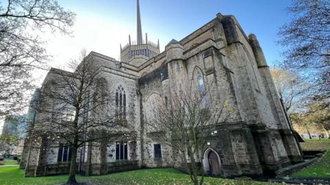 Blackburn Cathedral is a large imposing stone building set within a grassed area and surrounded by trees.