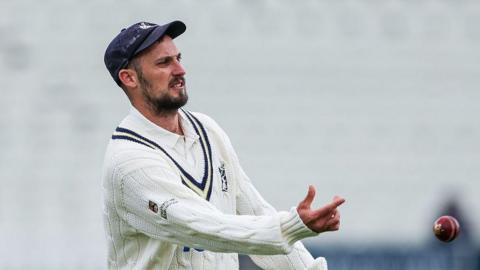 Ed Barnard wearing cricket whites and woollen longsleeve sweater plus club blue cap tossing ball to someone out of picture