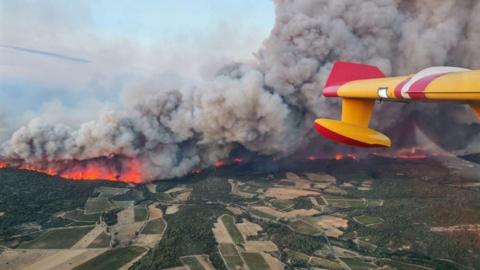 A wall of fire seen from an aircraft burns across the green patchwork of southern France fields.