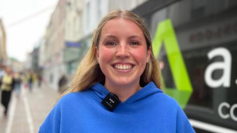 Eilidh is wearing a bright blue hoodie with the words “WHITE FOX” stands on a pedestrian shopping street. A small wireless microphone is clipped to the hoodie. Behind her, a shopfront window displays large white lettering and a green logo, while other pedestrians and buildings are visible in the softly blurred background.