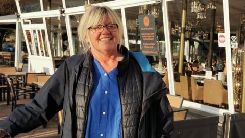 A smiling blonde woman with glasses wearing a blue shirt and a black jacket with tables and chairs behind her and a long window with more tables and chairs behind.