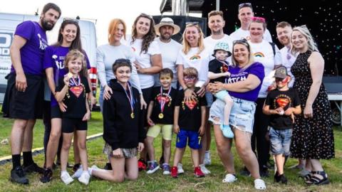 A large group photo of eleven adults and six children in front of them. They are standing outside in a field. Every child has a gold medal around their neck and some of them have had their face painted. It looks like they are at a fundraising event together. 