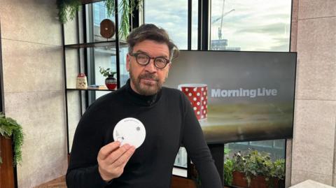 Nick Knowles in the Morning Live studio with a black jumper on holding up a smoke alarm