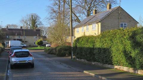 A residential road with cars parked next to the pavement on the left-hand side. To the right is a house made out of cream bricks and a long green bush. At the end of the road are two vans, greenery and another few houses.