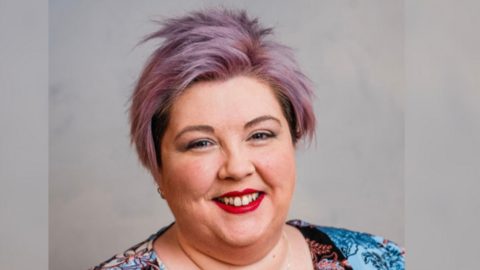 Ciara Hunter has short purple hair and is wearing makeup with red lipstick. She has a multicoloured top which is blue and black. The background is grey.