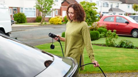 Woman wearing a green jumper is taking her charger to her car. She has curly brown hair. 