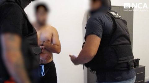 Two police officers in black vests arrest a shirtless man with handcuffs, his face blurred