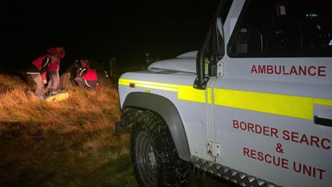 A rescue vehicle on a dark hillside shines its lights onto hill rescuers helping a casualty