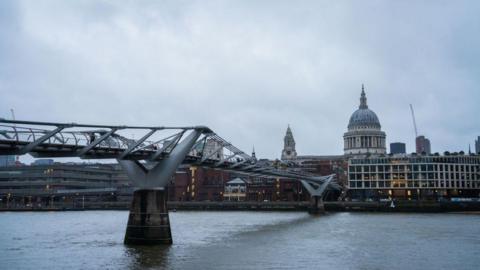 A view of the Millennium Bridge in London, spanning the River Thames under an overcast sky. The steel suspension bridge leads towards St. Paul's Cathedral, which stands prominently in the background.