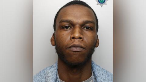 A mugshot of a man with short dark hair and wearing a blue shirt over a grey t-shirt.