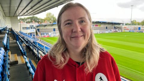 Jo Warillow in a red jumper, is in AFC Telford's stands, with the pitch behind her.