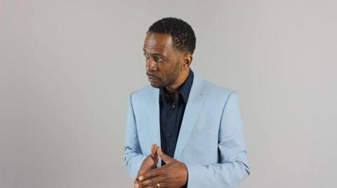 Granville Graham, wearing a light blue suit and navy shirt, in a studio head-and-shoulders photo in which he is looking slightly to the right. He has black hair and styled facial hair.
