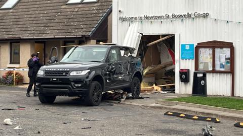 A black Range Rover with heavy damage to its side is pictured outside the Co-op in Newtown St Boswells after crashing into the shopfront, leaving debris scattered on the ground and a large hole in the wall. A police forensic officer wearing gloves photographs the scene as investigations into the attempted ram raid continue.
