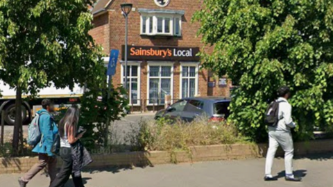 A shop with orange and brown Sainsbury's signage, with bushes in the foreground, and three people walking past.