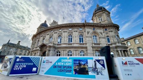 The exterior of the Hull Maritime Museum in Hull. The stone building has decorative carvings around the windows and has several towers which are topped by domes. The building is surrounded by a fence displaying advertising and branding.