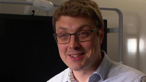 A man with glasses wearing a blue shirt standing in front of a TV in a hospital room.