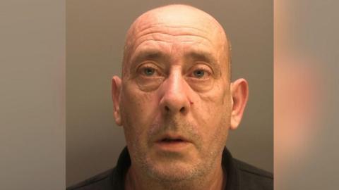 A police custody image of Mark Roberts, he has a bald head and stubble. He is wearing a black polo shirt and his mouth is slightly agape as he has his photo taken.