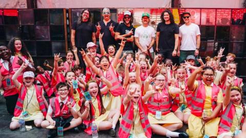 Group of schoolchildren smiling and waving wearing hi-vis jackets and sat on stage in front of the Foo Fighters' who are stood behind them.