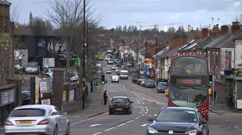 A view of a street in Stirchley. There are two lanes of traffic with cars and buses travelling on them. on the right are rows of shops and houses. On the left is a car park and people visible on the pavement in the background