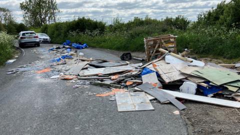 A large amount of litter spread out across the bend on a country lane. Cars are waiting to fit through the narrow gap.