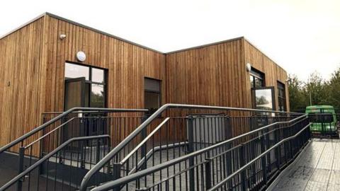 A single-storey building is clad with vertical timber. It has dark-framed glass doors and windows. It is raised up slightly with decking and a wheelchair-friendly entrance, surrounded by black railings.
