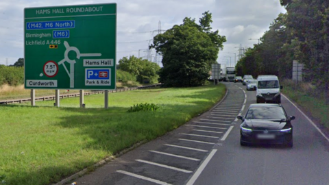 To the left is a large green road sign with the words Hams Hall Roundabout across the top. To the right is a lane of traffic, with vehicles heading towards the camera